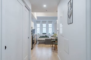 A narrow hallway leading to a bedroom with a bed, a desk, and a window.