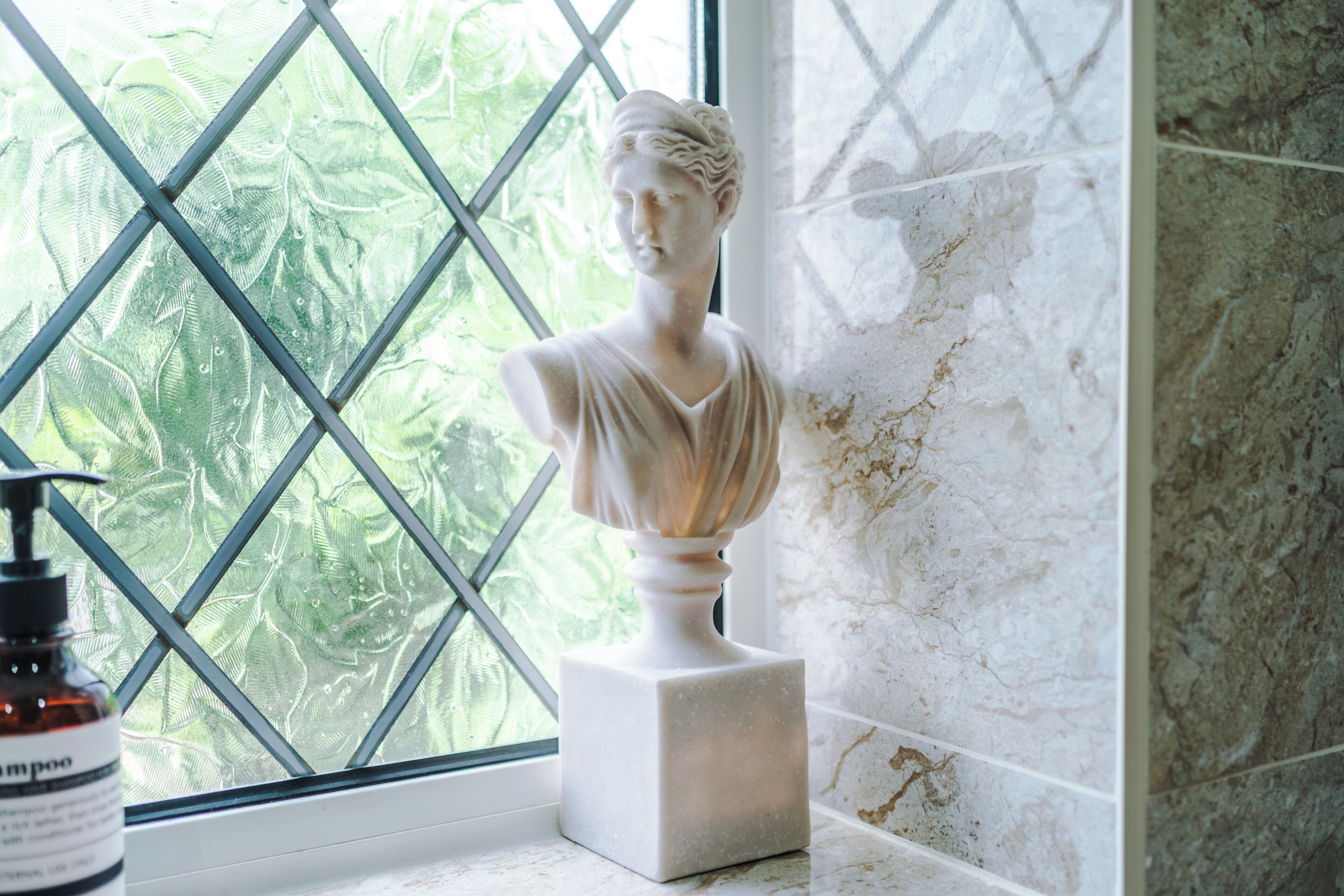 Una escultura en marbre de forma de bust sobre un pedestal, una ampolla de xampú i una finestra amb vista a vegetació.