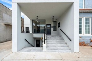 Una entrada moderna de edificio con escaleras, puerta de cristal y barandilla de metal.