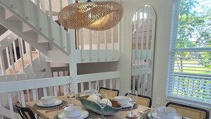 Comedor con una larga mesa dispuesta para cuatro, rodeada de sillas negras, una lámpara colgante tejida y una escalera al fondo.