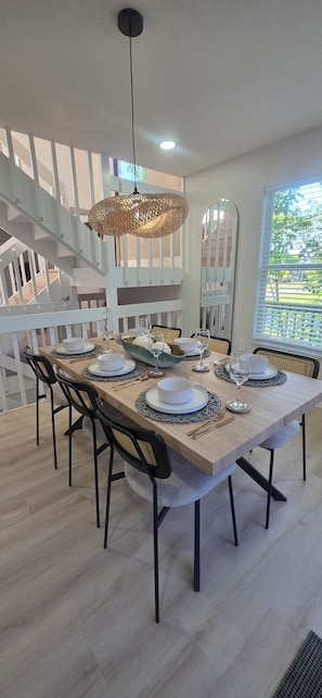 A dining area with a long table set for four, surrounded by black chairs, a woven pendant light, and a staircase in the background.