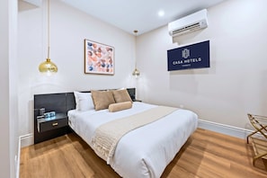 Habitación de hotel con cama, mesitas de noche, un reloj, aire acondicionado, una obra de arte enmarcada y un cartel con el nombre del hotel.