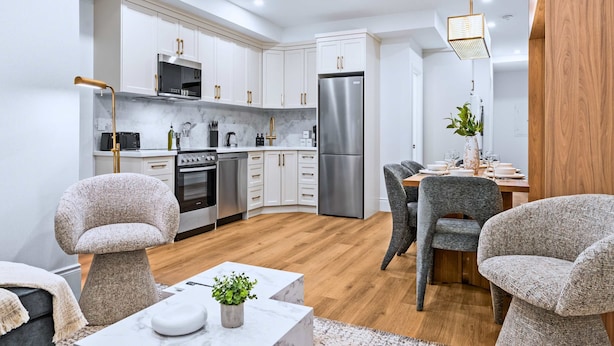 A modern kitchen with white cabinets, stainless steel appliances, and a dining area with grey upholstered chairs. - The Gerrard Group Basement CN Tower Transit Base (Toronto)