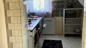 Et aflangt køkken med flisegulv, et køleskab, en vask og en hylde med genstande.