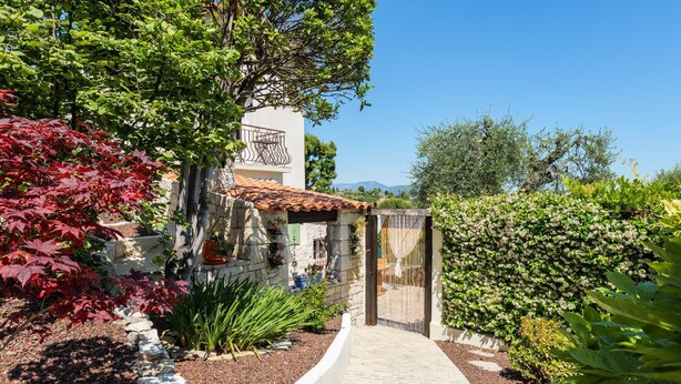 Un chemin menant à un bâtiment à la façade en pierre, entouré de différentes plantes et sous un ciel bleu clair.