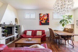 Ein Wohnzimmer mit einem roten Sofa, einem Holztisch, einem Esstisch mit Korbstühlen, einem Kamin und einem großen Gemälde an der Wand.
