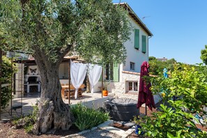 Ein Steinhaus mit einem großen Baum, Außenbereich mit Sitzgelegenheiten und Grillplatz.