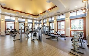 A spacious gym with various exercise machines, large windows, and a wooden floor.