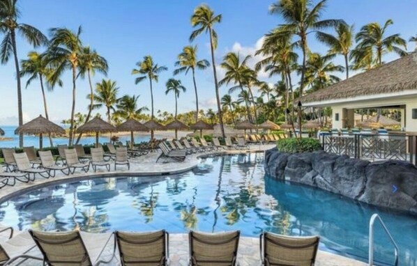 A pool area with lounge chairs, palm trees, and a building with a thatched roof.