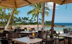 An outdoor dining area with tables and chairs, overlooking a beach and ocean.