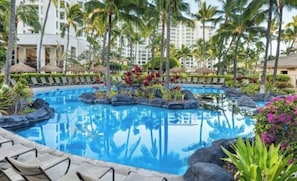 A hotel pool with lounge chairs, palm trees, and a reflection of the hotel buildings.