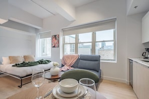 Ein modernes Wohnzimmer mit einer Couch, einem Esstisch mit Tellern und einem Glas sowie Blick auf die Stadt durch große Fenster.