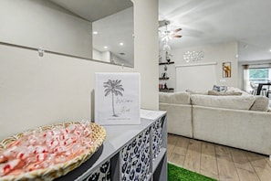 Ein modernes Wohnzimmer mit einem weißen Sofa, einem dekorativen Willkommensschild, einer Korb mit Snacks und einem Deckenventilator.