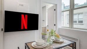 Une chambre moderne avec un téléviseur à écran plat fixé au mur, affichant le logo de Netflix, une table à manger avec de l’assiette blanche et un vase de fleurs, et une fenêtre donnant sur la ville.