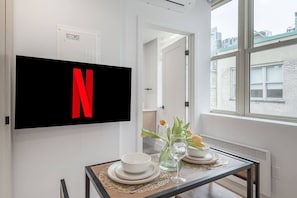 Ein modernes Zimmer mit einem wandmontierten Flachbildfernseher, der das Netflix-Logo anzeigt, einem Esstisch mit weißen Tellern und einer Blumenvase, sowie einem Fenster mit Blick auf die Stadt.