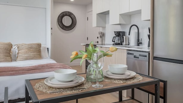 Une chambre d’hôtel moderne avec un lit, une petite table à manger avec des assiettes et un vase de fleurs, et un coin cuisine avec des placards et des appareils électroménagers.