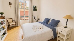 A bedroom with a bed, a chair, a nightstand, a lamp, a bookshelf, and a window with a view of buildings. - Charming Apt Steps from Eiffel Tower & Quai Branly (Paris)