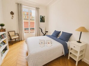 Ein Schlafzimmer mit Bett, Stuhl, Nachttisch, Lampe, Bücherregal und einem Fenster mit Blick auf Gebäude.