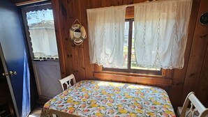 Quarto com mesa coberta por toalha de mesa estampada com frutas, cadeiras de madeira, janela com cortinas brancas e porta com moldura azul.
