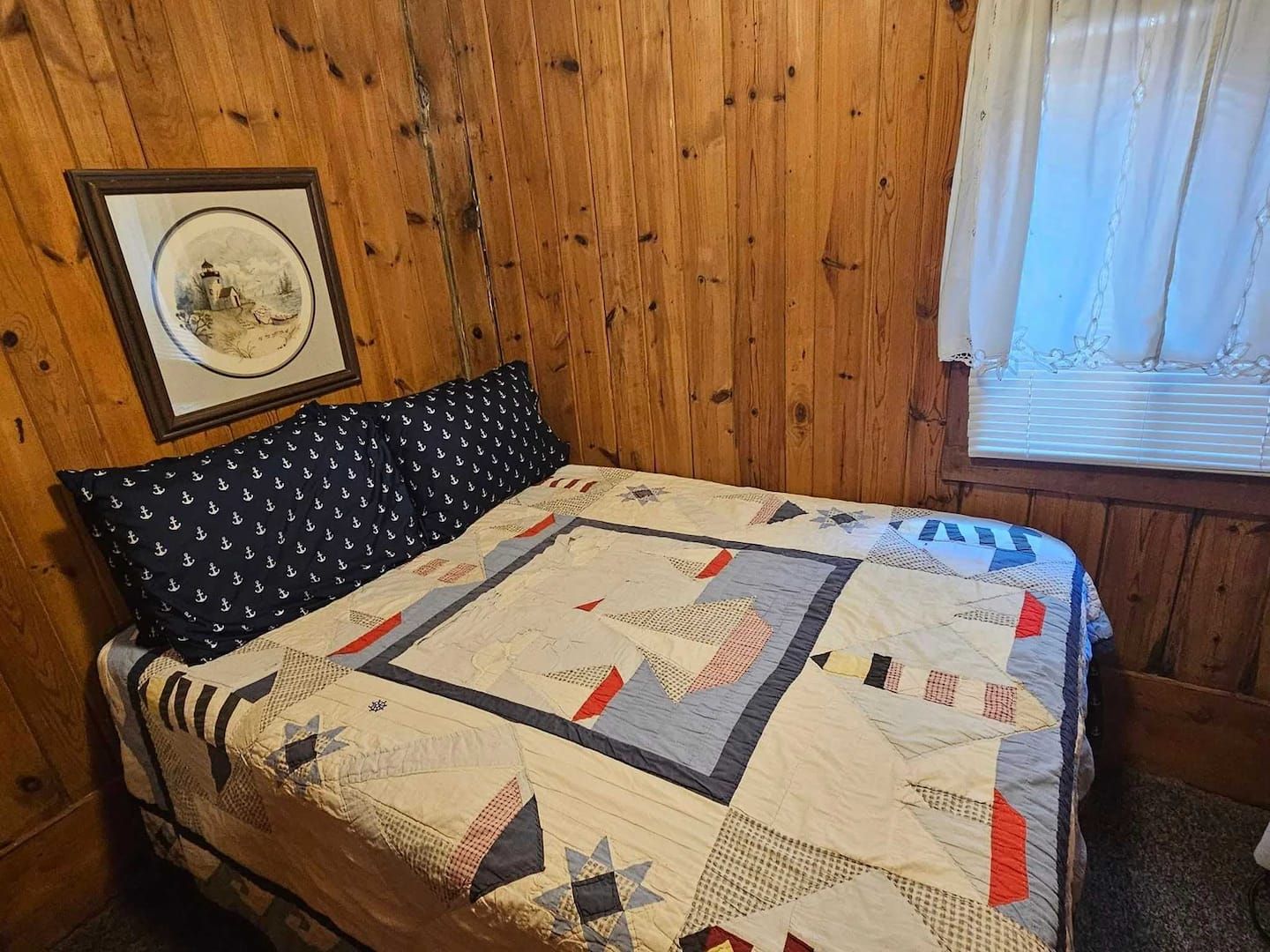 Une pièce aux murs en bois, un lit recouvert d’une courtepointe à motifs, un oreiller orné d’ancres et un tableau encadré accroché au mur.