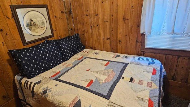 Quarto com paredes de madeira, cama com colcha estampada e travesseiro com desenhos de âncoras, além de um quadro na parede.