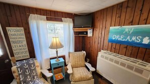Quarto com parede de painéis de madeira, televisão, mesa pequena com abajur, cadeira com tecido estampado com padrão de âncora e um letreiro com a inscrição 'My Dreams'.