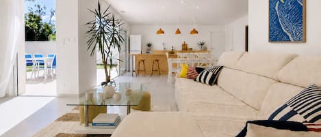 A modern living room with a sofa, a glass coffee table, and a dining area with chairs and a kitchen in the background.