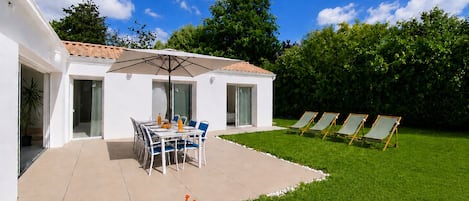 A modern house with a patio, a dining table with chairs, a covered outdoor area, and a lawn with sun loungers.