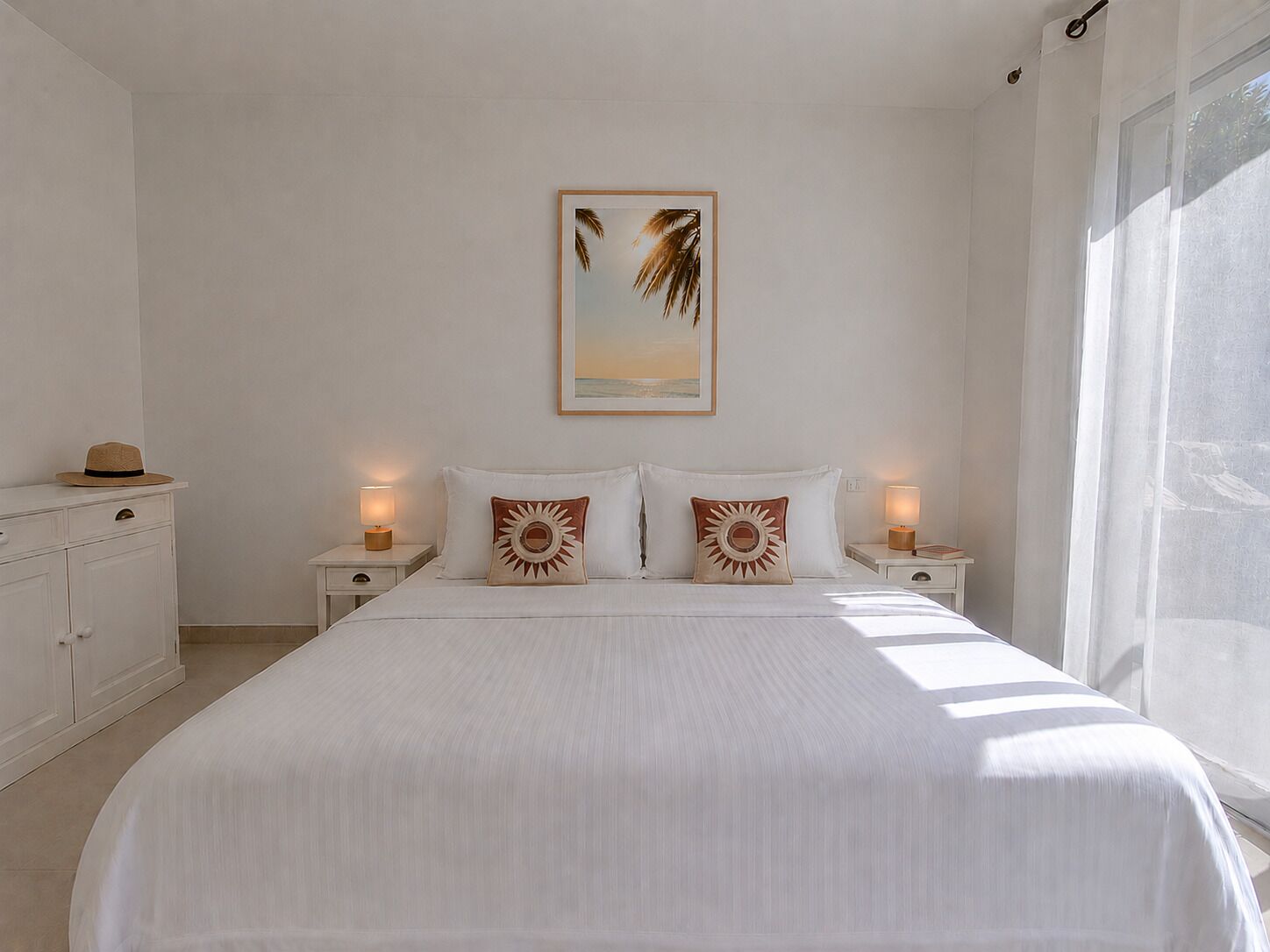 A bedroom with a large bed, two bedside tables with lamps, a framed picture on the wall, and a window with sheer curtains.