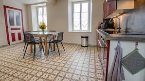 A kitchen with a tiled floor, a dining table with chairs, a trash bin, and a window. - Le Nid d'Hirondelles (FONTAINE LES GRES)