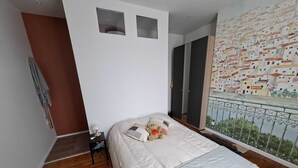 Quarto com cama, mesa de cabeceira, cômoda, janela com vista para edifícios e porta.