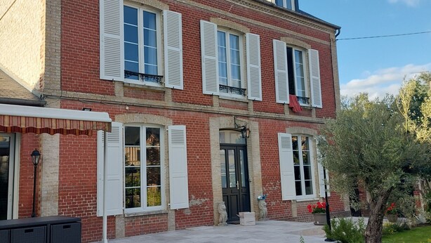Uma casa de dois andares de tijolos com venezianas brancas, uma porta da frente e um pequeno jardim com uma árvore e flores.