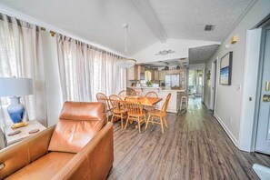 Sala de estar con sofá de cuero, mesa de comedor con sillas, zona de cocina y pasillo que conduce a una puerta.