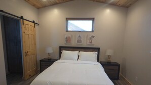 Un dormitorio con techo de madera, una cama con ropa blanca, dos mesitas de noche con lámparas, una puerta de madera y una ventana.