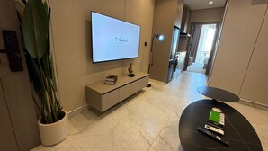 A modern living room with a flat-screen TV, a potted plant, a small table with a remote control, and a hallway leading to a kitchen area. - 401 Tara-jre Jabal Al Lweibdeh With Stunning View (Amman)