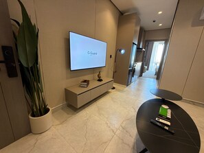 Uma sala de estar moderna com televisão de tela plana, uma planta em vaso, uma mesinha com um controle remoto e um corredor que dá para uma cozinha.