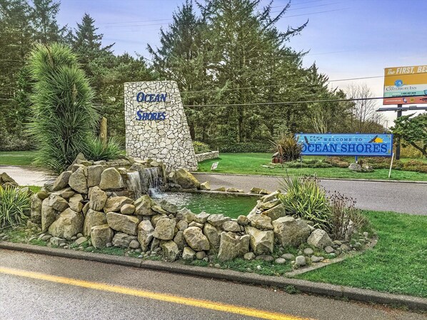 A stone sign with 'Ocean Shores' written on it, a small waterfall feature, and a road with a yellow line. - Ground Floor 2-Bedroom Suite - Pet Friendly! (Ocean Shores)
