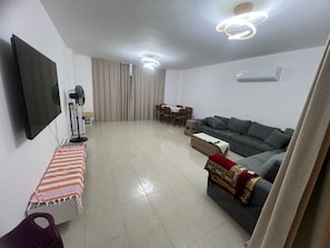 A living room with a flat-screen TV, a grey sofa, a dining table with chairs, a bench, and a ceiling fan.