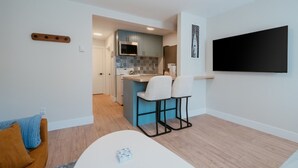 Een moderne keuken met een magnetron, kasten en een klein kookeiland met barstoelen. Er is een eethoek met een tafel en stoelen, en een aan de muur bevestigde televisie.