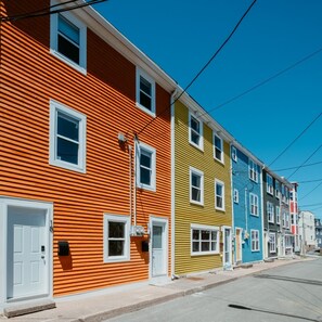 A row of colorful row houses with white doors and windows, a clear sky, and a visible street. - The Orange Bean | 1 Bed 1 Bath (St. John`s)