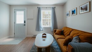 Een kamer met een witte deur, een raam met gordijnen, een bruine bank, een kleine tafel, en lijstwerken aan de muur.