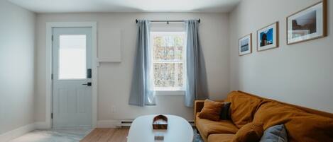 Een kamer met een witte deur, een raam met gordijnen, een bruine bank, een kleine tafel, en lijstwerken aan de muur.
