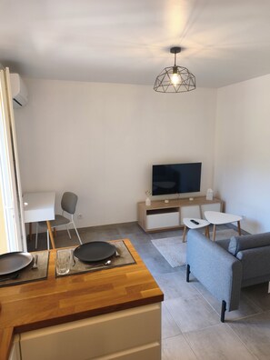 A modern living room with a sofa, a dining table, a TV, and a ceiling light fixture. - 2 People | Near town center and beaches | Air-conditioned | Garden (Cavalaire sur Mer)