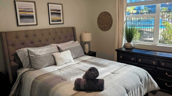 Een slaapkamer met een gebobbelde hoofdeinde, een bed met gestreept beddengoed, een bedlamp, een decoratief wandobject en een uitzicht op een zwembad door een raam.