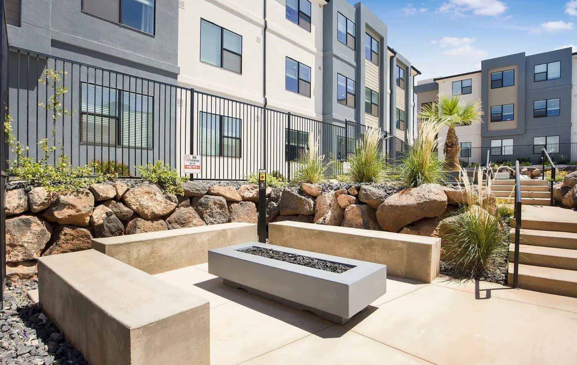 A modern outdoor seating area with a fire feature, stone walls, and a staircase leading to a building with multiple windows.