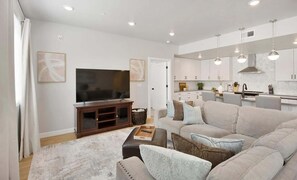 Uma sala de estar moderna com televisão de tela plana, sofá, mesa de centro e área de cozinha com luminárias pendentes.