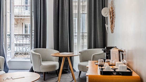 Ein modernes Hotelzimmer mit Blick auf die Stadt, zwei gemusterte Sessel, ein runder Holztisch und eine Ablage mit Kaffeezubehör.