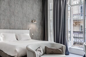 A bedroom with a large bed, a sofa, a window with curtains, and a wall-mounted light fixture. - Prestige Suite & Balcony (Bordeaux)