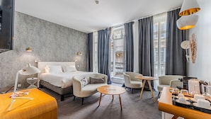 Ein modernes Hotelzimmer mit einem Bett, zwei Sesseln, einem kleinen runden Tisch, einer Kaffeestation und Blick auf die Stadt durch große Fenster.