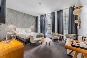 A modern hotel room with a bed, two armchairs, a small round table, a coffee station, and a view of the city through large windows. - Prestige Suite & Balcony (Bordeaux)